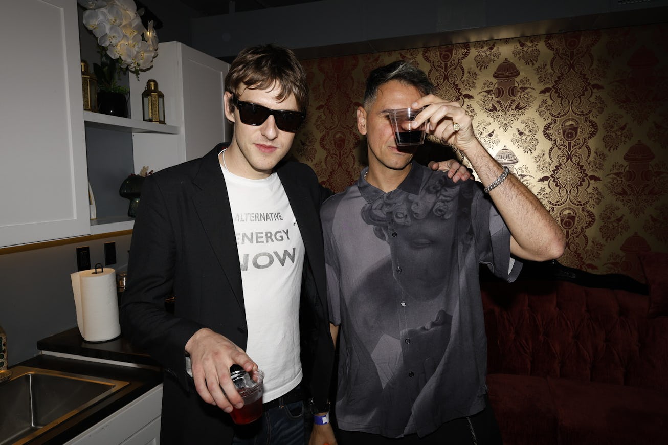 Two men stand together in a stylish room, holding drinks. One wears sunglasses and a black blazer ov...
