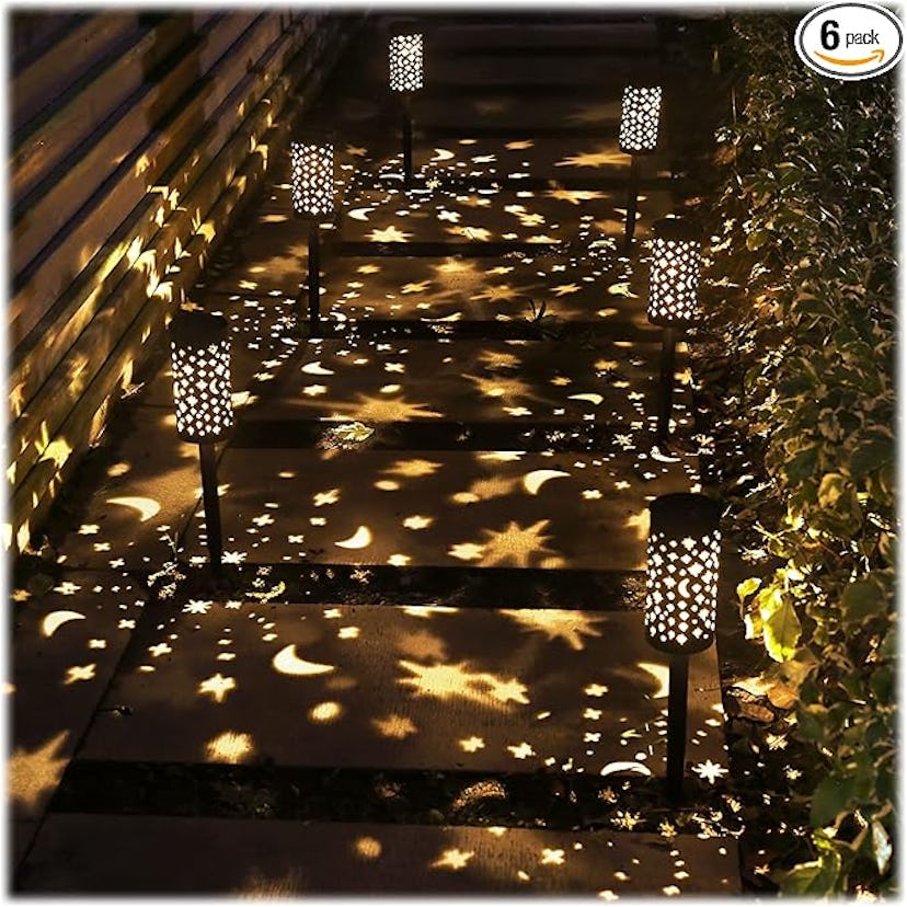 A walkway illuminated by decorative lanterns casting star and moon patterns on the ground, surrounde...
