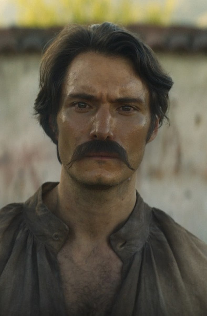 A man in a historical costume stands solemnly, with dark hair and a prominent mustache, in front of ...