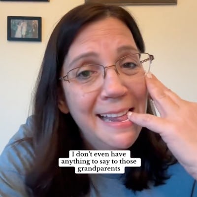 A woman with glasses smiles while speaking on the phone. She gestures expressively with her hand, sitting in a room with framed artwork on the wall.