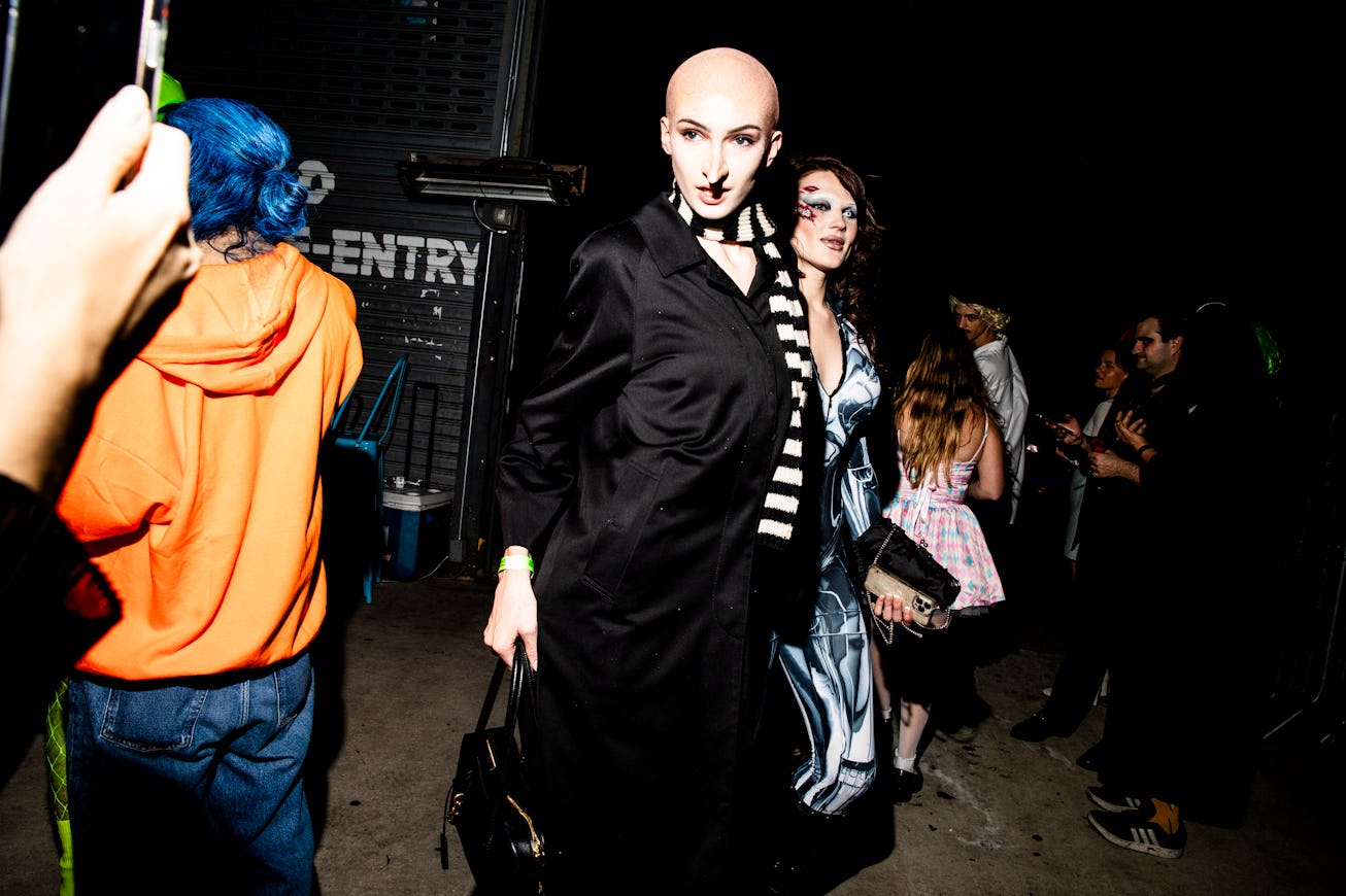 A group of diverse individuals in stylish, bold outfits stands in a dimly lit venue. One person with...