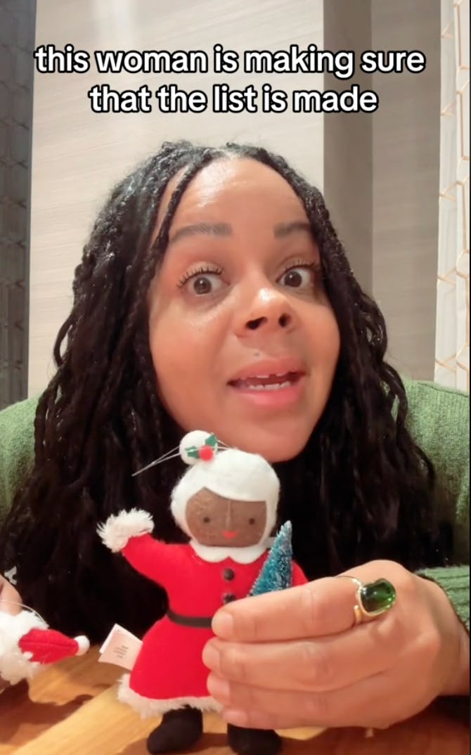 A woman with long, curly hair is animatedly speaking while holding a handmade doll in a red dress and a small blue tree, with a playful expression.