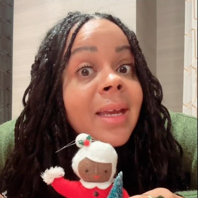 A woman with long, curly hair is animatedly speaking while holding a handmade doll in a red dress and a small blue tree, with a playful expression.