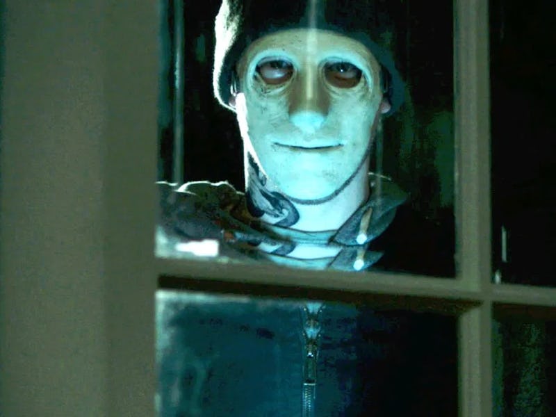 A figure with a pale mask stands outside a window, illuminated by eerie light, creating a tense and ...