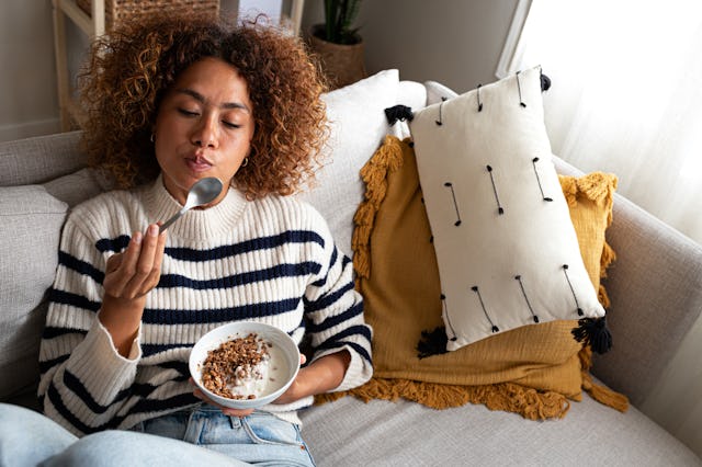 A woman with curly hair in a striped sweater enjoys a bowl of yogurt on a cozy couch, surrounded by ...