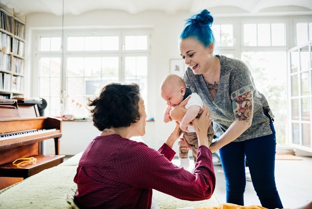 A smiling woman with blue hair holds a baby, while another woman with curly hair joyfully reaches to...