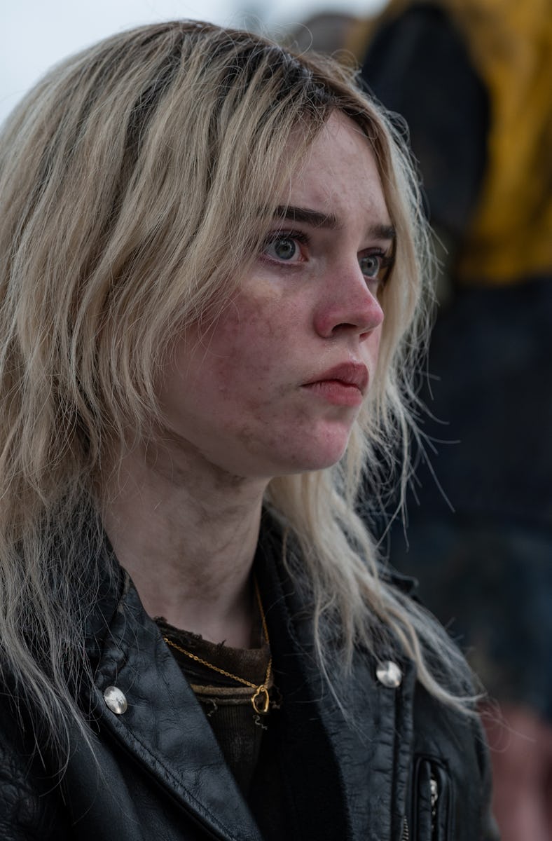 A young woman with unkempt blonde hair and a serious expression sits in the foreground, wearing a bl...