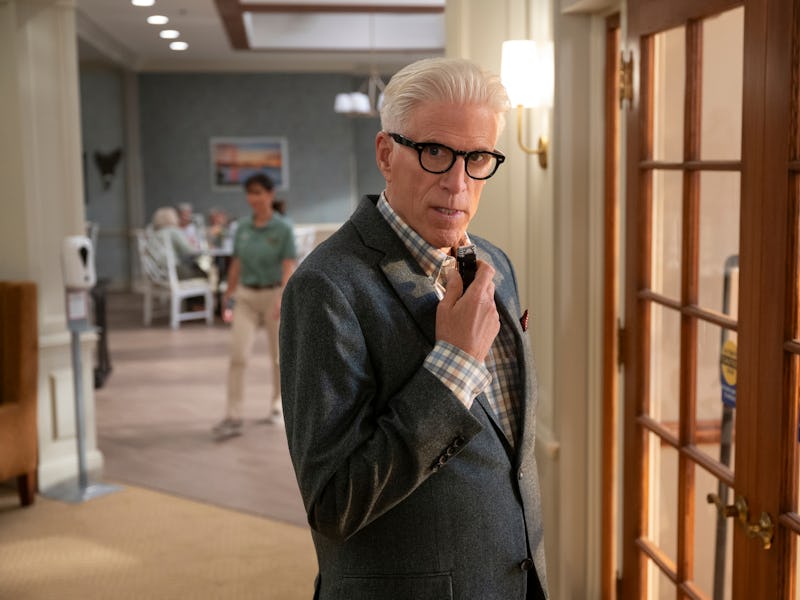 A man with slicked-back silver hair and glasses stands near a glass door, giving a thoughtful look w...