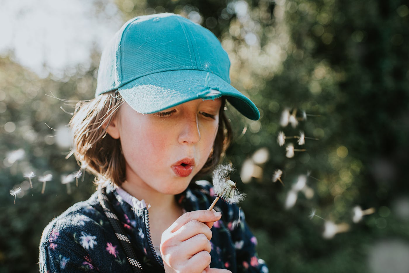 A young child wearing a turquoise cap blows softly on a dandelion, sending its seeds drifting into the air, surrounded by greenery.