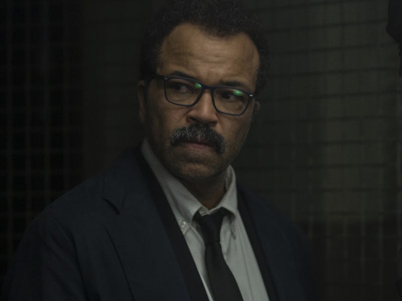 A man in a suit and glasses stands with a serious expression, looking to the side, while a dark figu...
