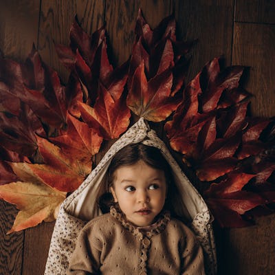 A child lies on a wooden floor, surrounded by vibrant autumn leaves in shades of red and orange, creating a whimsical and cozy atmosphere.