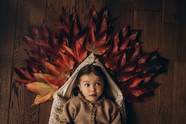 A child lies on a wooden floor, surrounded by vibrant autumn leaves in shades of red and orange, cre...