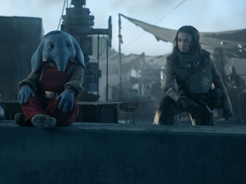 A whimsical character with an elephant-like head sits on a ledge, while a woman in a rugged outfit o...