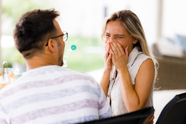 A couple is seated at a table outdoors, laughing together. The woman playfully covers her face while...