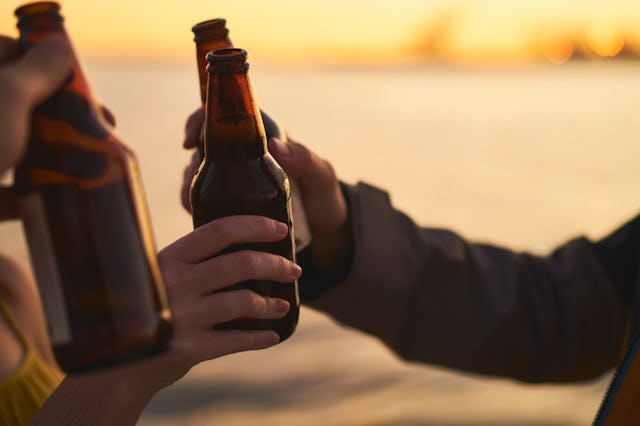 Three hands holding beer bottles in a toast against a sunset backdrop. The serene water and warm col...