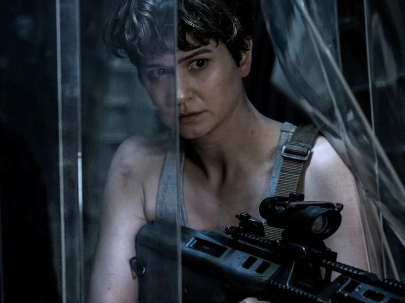 A woman with short hair, wearing a sleeveless top, grips a rifle while peering through a transparent...