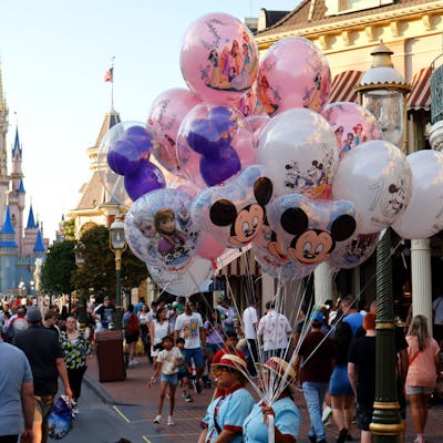 A bustling theme park scene featuring people walking down a pathway, surrounded by colorful balloons, with a fairytale castle in the background.