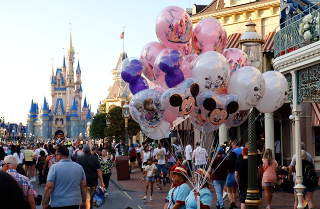 A bustling theme park scene featuring people walking down a pathway, surrounded by colorful balloons...