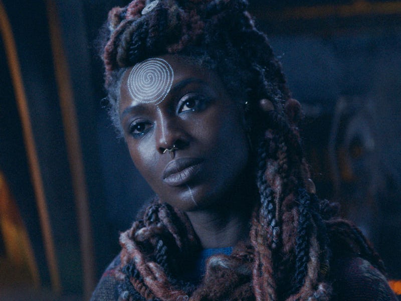 A close-up of a woman with intricate, dark braided hair and a decorative forehead emblem. She has a ...