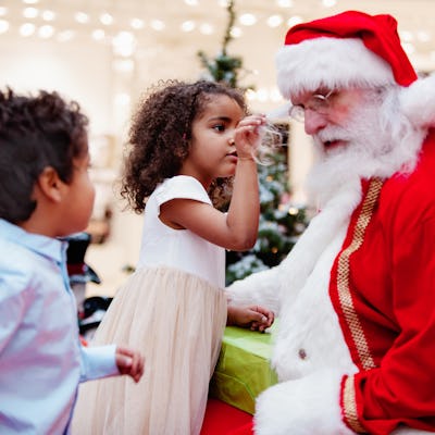 A young girl in a white dress playfully touches Santa's beard while a boy in a blue shirt watches. Festive decorations are visible in the background.
