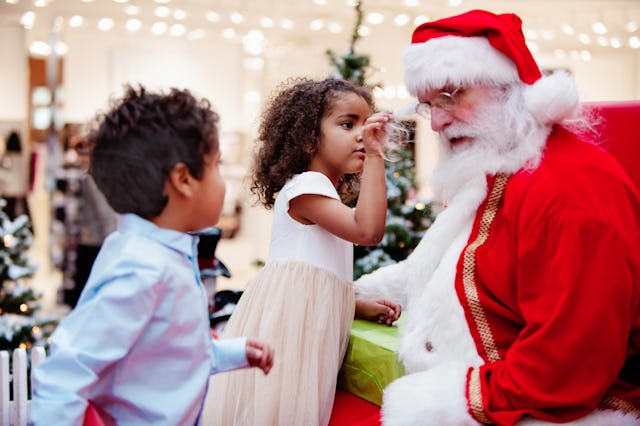 A young girl in a white dress playfully touches Santa’s beard while a boy in a blue shirt watches. F...