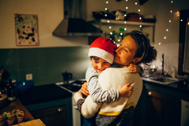 A joyful moment in a cozy kitchen, a mother hugs her smiling child wearing a Santa hat, festive deco...