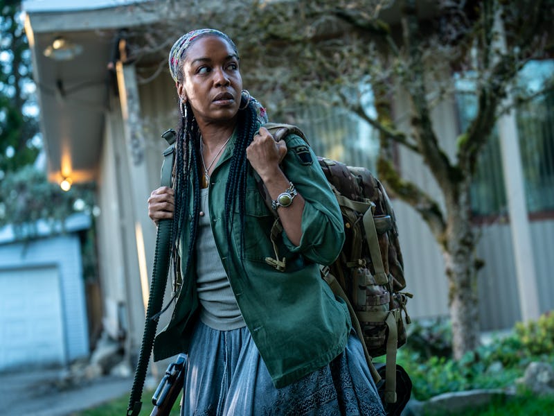 A woman stands outdoors, carrying a backpack and a firearm. She wears a green jacket and has braided...
