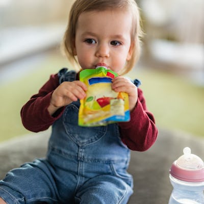 A young child sits on a soft surface, eating from a colorful pouch while holding a baby bottle nearby. They wear denim overalls and a red top.