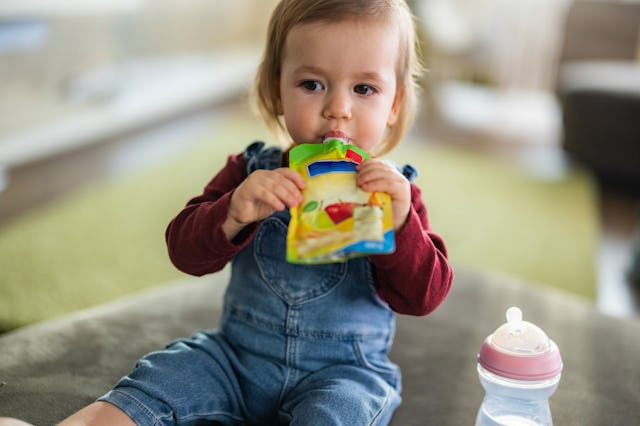 A young child sits on a soft surface, eating from a colorful pouch while holding a baby bottle nearb...