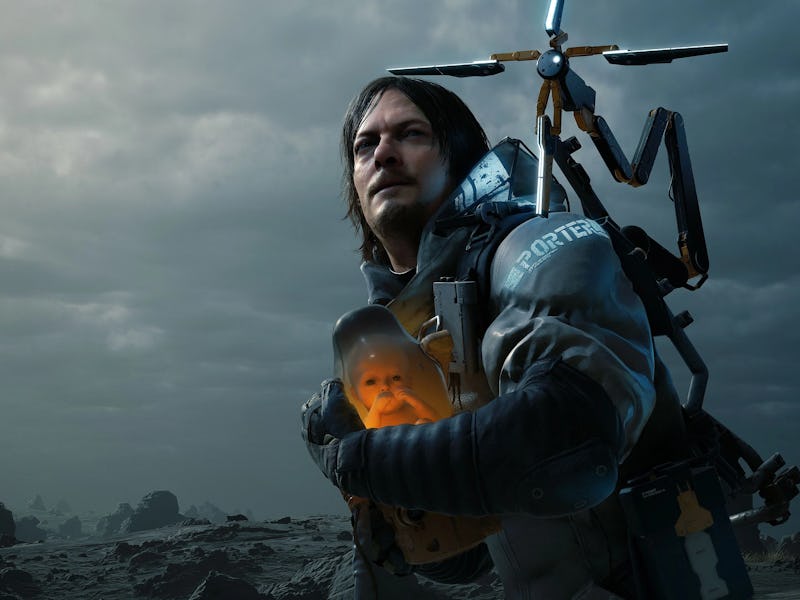 A rugged man stands in a desolate landscape, holding a glowing infant figure. He wears a high-tech s...