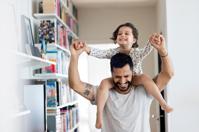A smiling child sits on a man's shoulders, both holding hands in joy. They are indoors, surrounded b...
