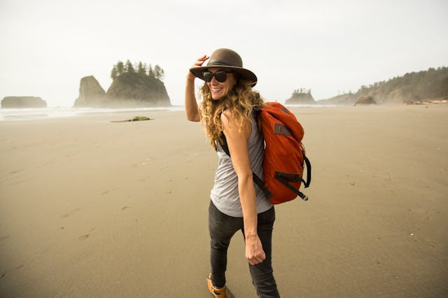 A woman with long hair, wearing a hat and sunglasses, smiles while walking on a beach with a backpac...