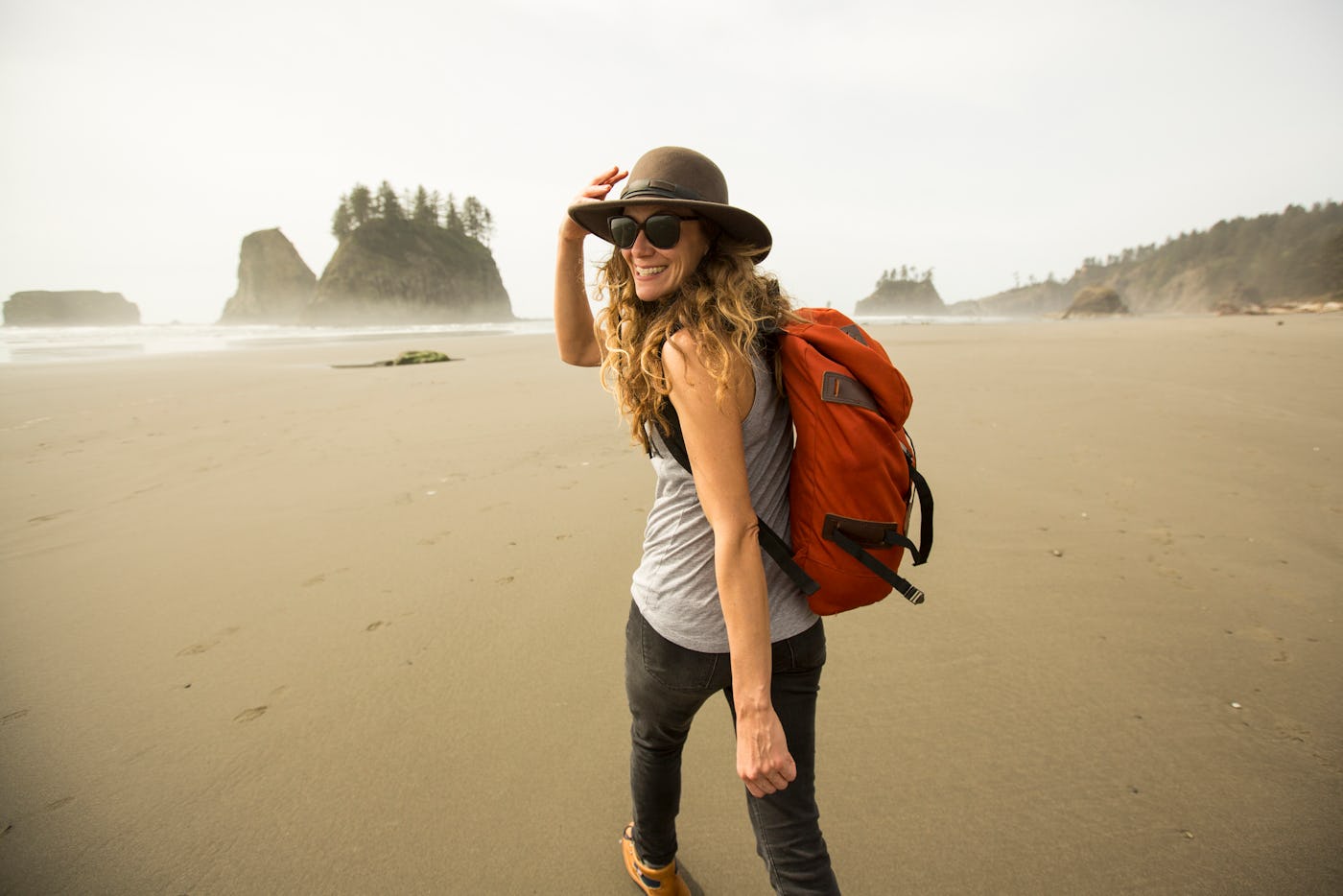 A woman with long hair, wearing a hat and sunglasses, smiles while walking on a beach with a backpack. Scenic rocky formations are in the background.