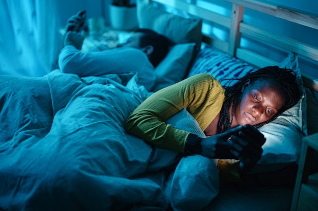 A woman in bed using her phone at night, illuminated by its glow, while a man lies beside her, also ...