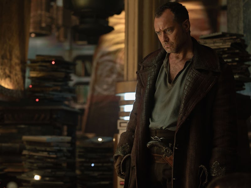 A man in a dark, worn coat stands thoughtfully in a dimly lit, book-filled room. Shelves are stacked...