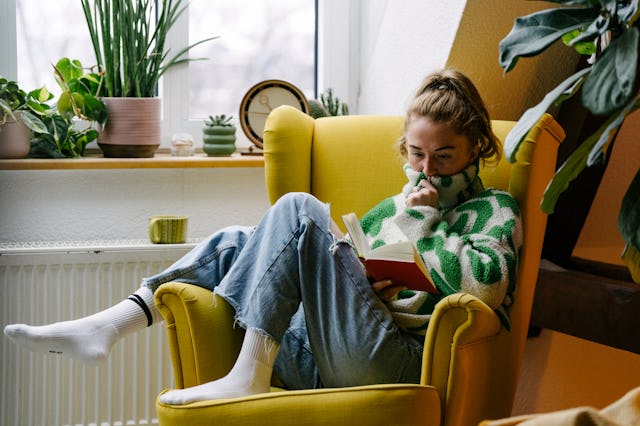 A woman sits comfortably in a bright yellow armchair, reading a book while wrapped in a cozy green s...