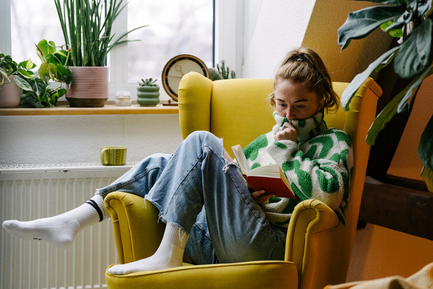 A woman sits comfortably in a bright yellow armchair, reading a book while wrapped in a cozy green sweater. Indoor plants decorate the space.