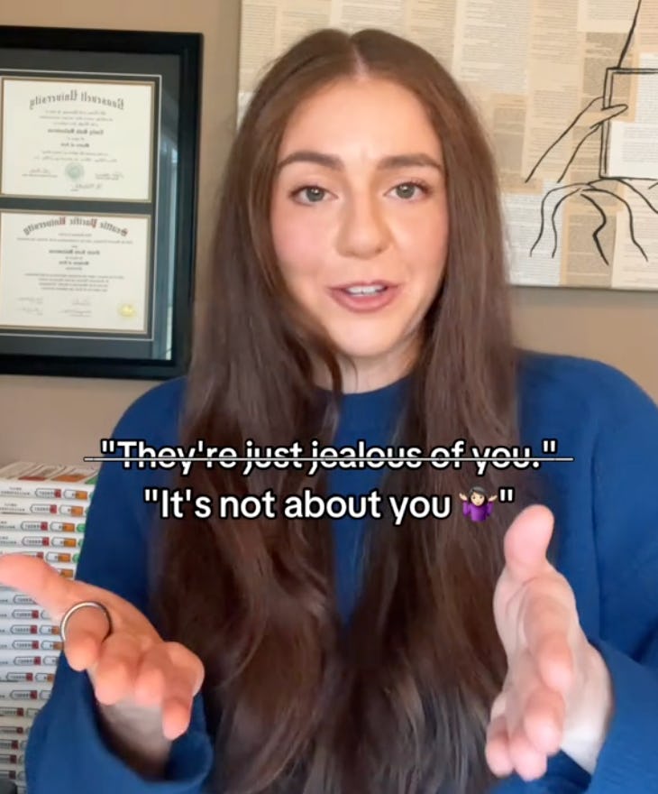 A person with long brown hair gestures while speaking. The background features framed certificates and a stack of books. Text overlay includes phrases about jealousy and self-focus.