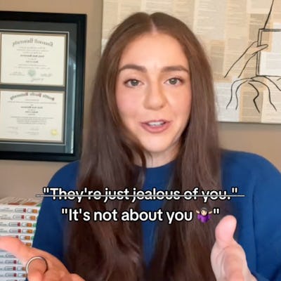 A person with long brown hair gestures while speaking. The background features framed certificates and a stack of books. Text overlay includes phrases about jealousy and self-focus.