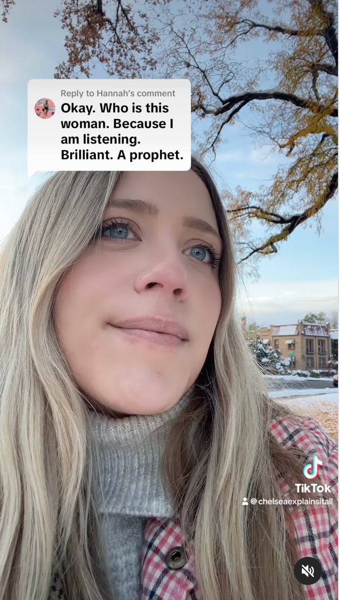 A close-up of a woman with long, light hair, wearing a cozy sweater and a plaid coat, outdoors with snow and trees in the background.