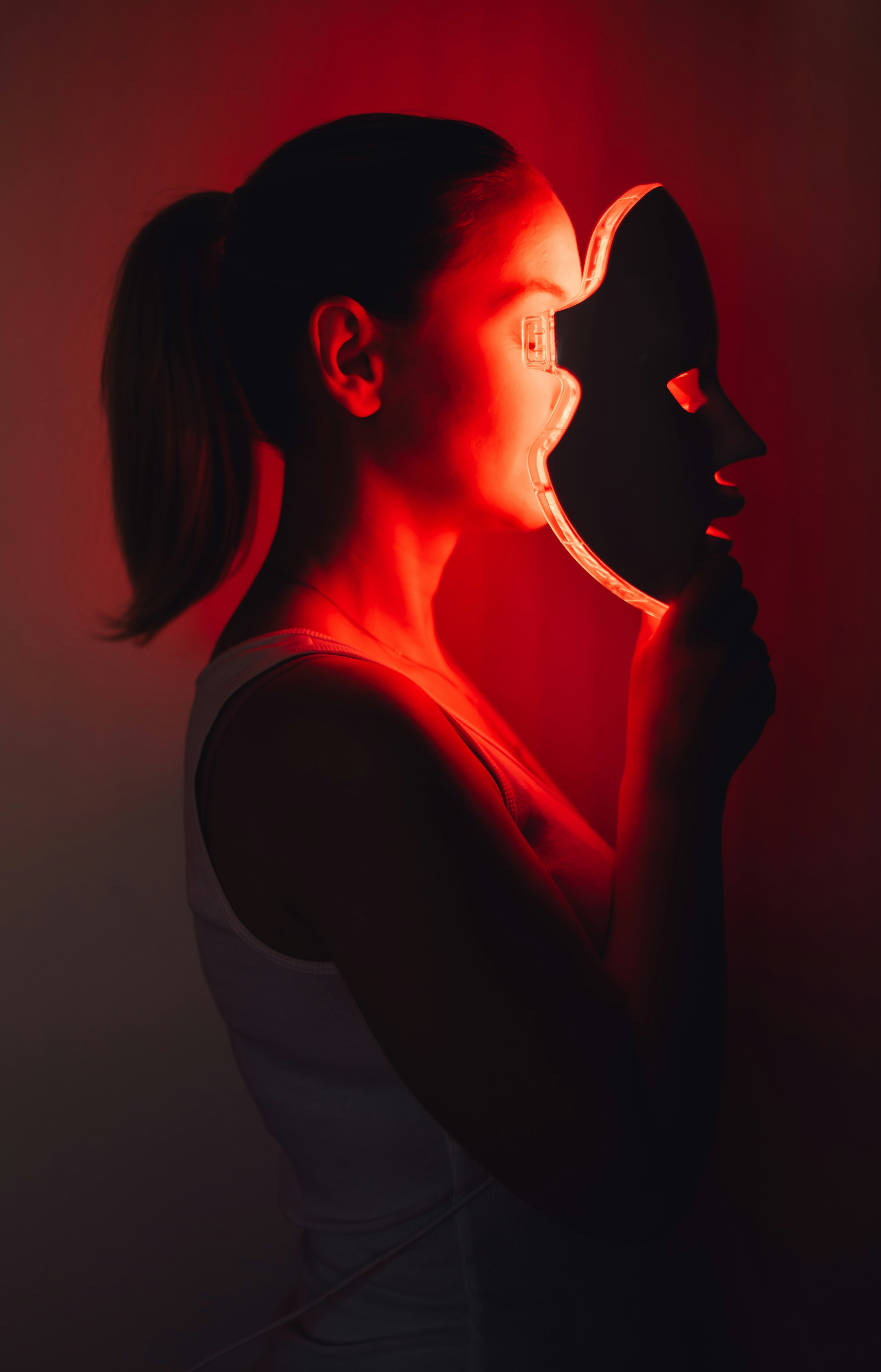 A woman holds a red light LED mask in front of her face