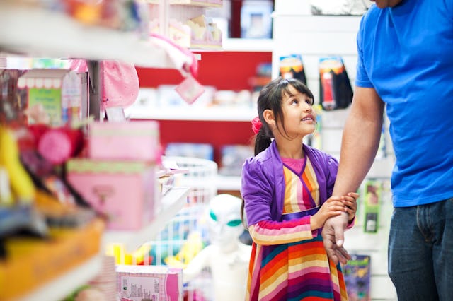 A young girl in a colorful striped dress and purple jacket smiles as she holds hands with an adult i...