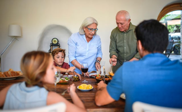A family shares a meal at a dining table, with two elders serving food. Children and adults enjoy th...