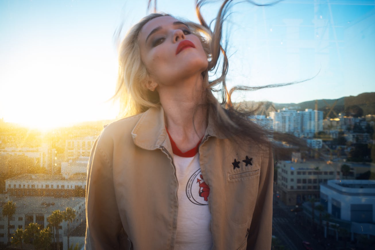 A young woman with long, flowing hair stands against a sunset backdrop, wearing a light jacket over ...