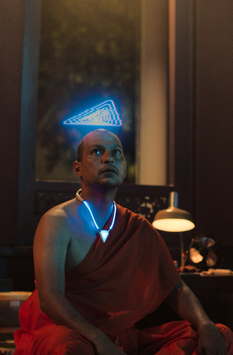 A monk sits in a dimly lit room, wearing traditional robes. A soft blue geometric light hovers above...