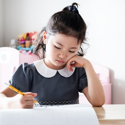 A young girl in a gray dress with a white collar sits at a wooden table, thoughtfully writing on paper. She has curly hair and a focused expression.