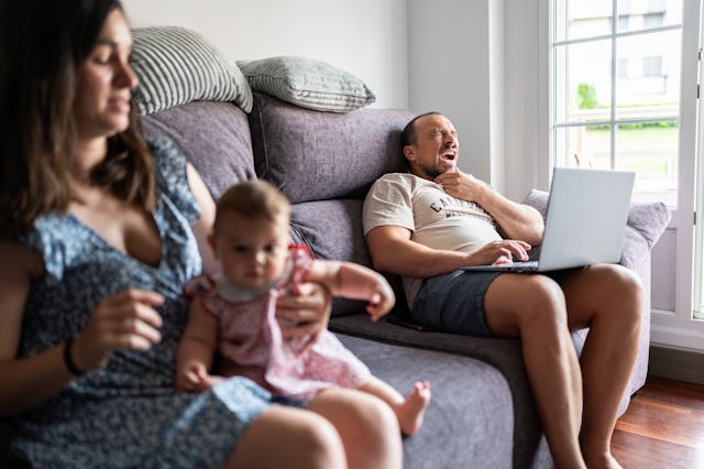 A woman holds a baby while sitting beside a man on a couch. He appears tired and works on a laptop, ...