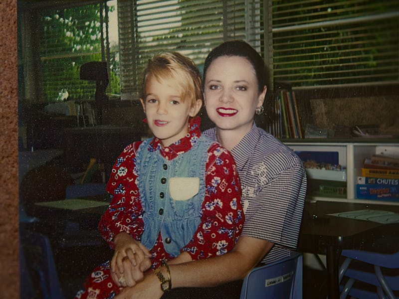 A smiling woman sits with a young girl on her lap, both dressed in colorful outfits. The background ...