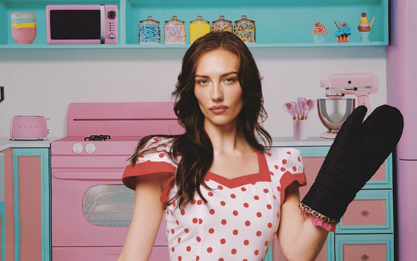 A confident woman in a polka dot dress stands in a colorful kitchen, wearing an oven mitt and surrou...
