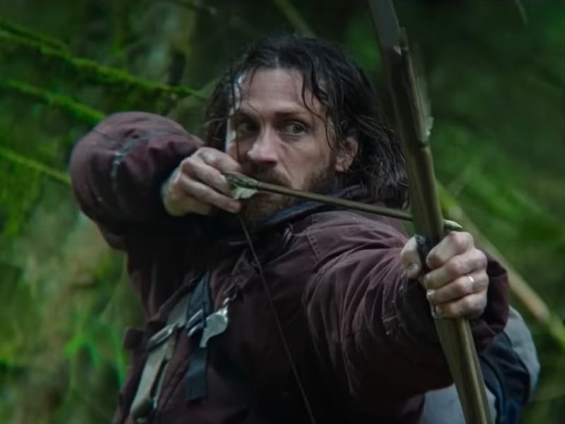 A rugged man is aiming an arrow with a bow in a dense, green forest, showcasing tension and focus in...